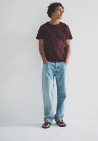 Bordeauxrode katoenen T-shirt, lichtblauwe spijkerbroek en zwarte slippers. Model staat met handen in de zakken tegen een witte muur.