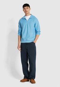 Lichtblauwe ritssluiting pullover met geribbelde manchetten, gecombineerd met donkerblauwe broeken en bruine loafers. Zichtbaar logo op de borst.