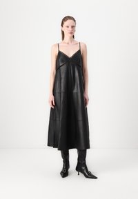 Schwarzes Leder-Midi-Kleid mit dünnen Trägern, einem V-Ausschnitt und dekorativen Ausschnitten. Kombiniert mit spitzen schwarzen Ankle-Boots. Gesamt glatte Textur.