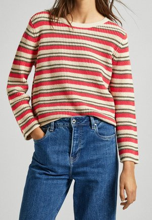 Pull en tricot avec des rayures horizontales crème, rouge et olive, porté avec un jean en denim bleu, main dans la poche.