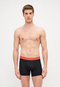 Black boxer shorts with a red elastic waistband featuring the brand logo. Smooth texture and fitted design for comfort and support.