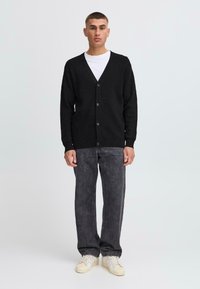 Black wool-blend cardigan with a V-neck, front buttons, and ribbed cuffs, paired with dark gray jeans and beige footwear.