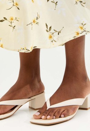 Pies de piel oscura usando sandalias tipo chancla con tacón cuadrado beige bajo una falda crema con estampado de pequeñas flores amarillas.