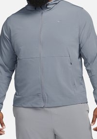 Lichte, grijze Nike zip-up jas met een capuchon, elastische manchetten, zijzakken en een gladde textuur, met een klein logo op de borst.