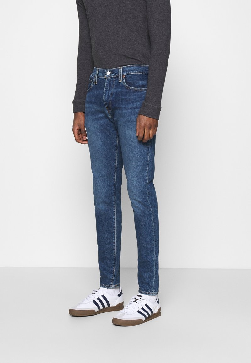 Personne portant un jean slim bleu, des baskets blanches avec des bandes noires et un haut gris foncé à manches longues, debout devant un fond uni.