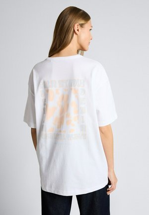 Frau mit langen Haaren, die ein lockeres weißes T-Shirt mit einem hellbeigen grafischen Druck auf dem Rücken trägt, kombiniert mit dunklen Hosen, seitlich stehend.