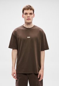 Brązowa bawełniana koszulka z okrągłym dekoltem, krótkimi rękawami, luźnym krojem, z małą białą naszywką z logo na środku.