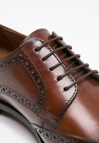 Scarpa da dress in pelle marrone con dettagli perforati, design con lacci, finitura lucida, punta arrotondata e suola nera a contrasto.