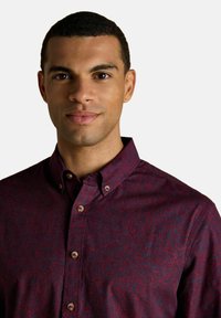 Jauns vīrietis ar īsiem melniem matiem, ģērbies tumši sarkanā rakstainā krekla ar pogām, skatās tieši kamerā ar neitrālu izteiksmi.