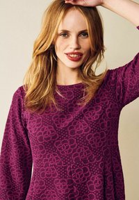 Violetti mekko, jossa on kuvioitu kukkakuvio, pyöreä pääntie ja pitkät hihat. Kangas vaikuttaa pehmeältä ja helma levenee alaspäin.