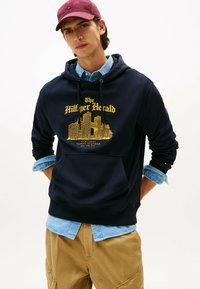 Marineblaue Kapuzenjacke mit gelbem Grafikdruck, der "The Hilfiger Herald" zeigt, und Kordeln. Kombiniert mit einem hellblauen Denim-Hemd und khakifarbenen Hosen.