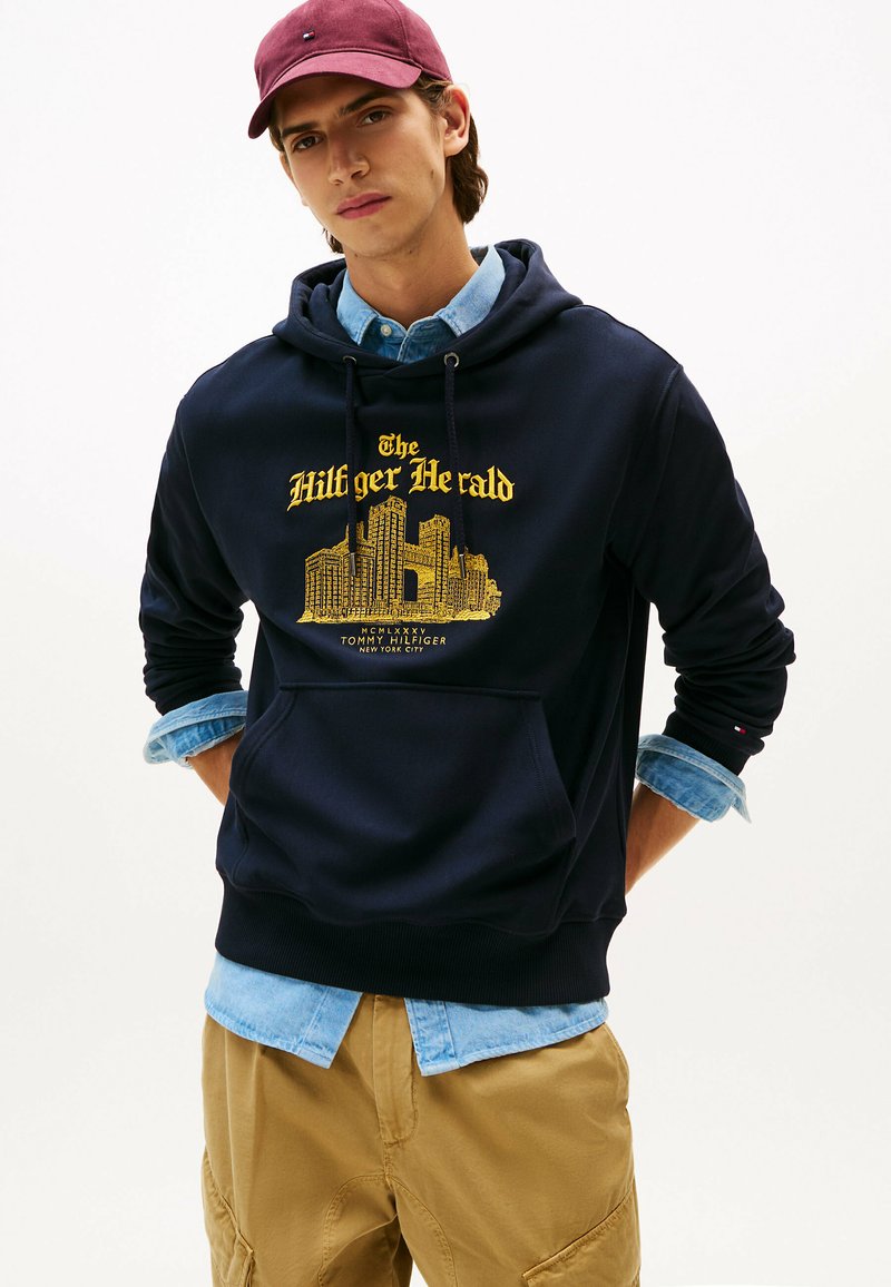Marineblaue Kapuzenjacke mit gelbem Grafikdruck, der "The Hilfiger Herald" zeigt, und Kordeln. Kombiniert mit einem hellblauen Denim-Hemd und khakifarbenen Hosen.