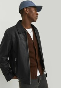 Black leather jacket with a collared design, side zipper pockets, layered over a brown cardigan and white t-shirt, paired with a blue cap.