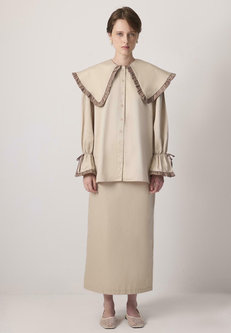 Νεαρή γυναίκα που στέκεται φορώντας μπεζ πουκάμισο με κουμπιά και υπερμεγέθη γιακά, μαζί με μακρύ φούστα στο ίδιο χρώμα, και πλεκτά παπούτσια, σε απλό φόντο.