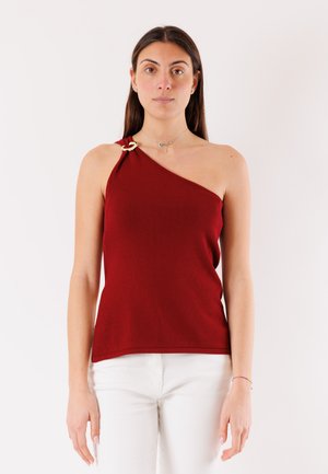 Donna che indossa una maglia rossa asimmetrica senza maniche con una fibbia dorata su una spalla, abbinata a pantaloni bianchi, in piedi davanti a uno sfondo neutro.