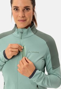 Helle grüne Zip-Jacke aus strukturiertem Stoff, mit grauen Akzenten, Stehkragen und Reißverschluss-Tasche. Auf der Vorderseite befindet sich das "VAUDE"-Logo.