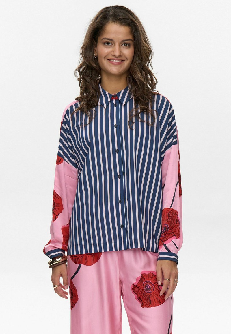 Chemise à rayures avec des rayures verticales bleu marine et blanches, manches roses ornées de grands motifs floraux rouges, et une coupe décontractée.