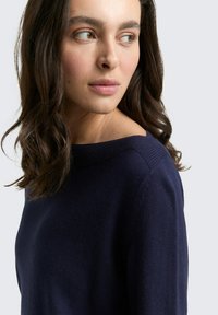 Frau mit welligem, dunklem Haar, die einen marineblauen Strickpullover trägt, seitlich steht und nach links schaut, vor einem schlichten hellen Hintergrund.