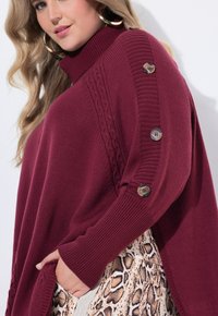 Un pull en maille bordeaux avec un col haut, une texture côtelée et des accents de boutons sur les manches ; porté par-dessus un vêtement clair à motifs.