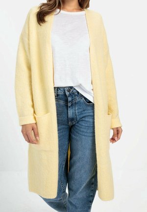 Femme portant un long cardigan jaune doux par-dessus un t-shirt blanc et un jean bleu, debout devant un fond blanc.