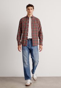 Geruit overhemd in rood, groen en crèmekleurig, met een button-down kraag, gecombineerd met lichtblauwe spijkerbroek en witte sneakers met bruine accenten.