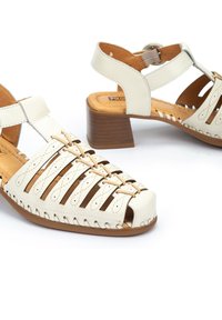 White leather sandals with an open-toe design, featuring a woven upper, cut-out sections, and a brown block heel.