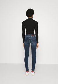 Jeans ajustados de mezclilla azul con bolsillos traseros, un parche de cuero en la cintura y una textura suave. Combinados con zapatillas blancas y una camiseta negra.