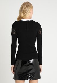 Černý žebrovaný top s dlouhým rukávem a krajkovými detaily na ramenou, spárovaný s lesklou černou mini sukní. Obsahuje zapínání na zip na zadní straně.