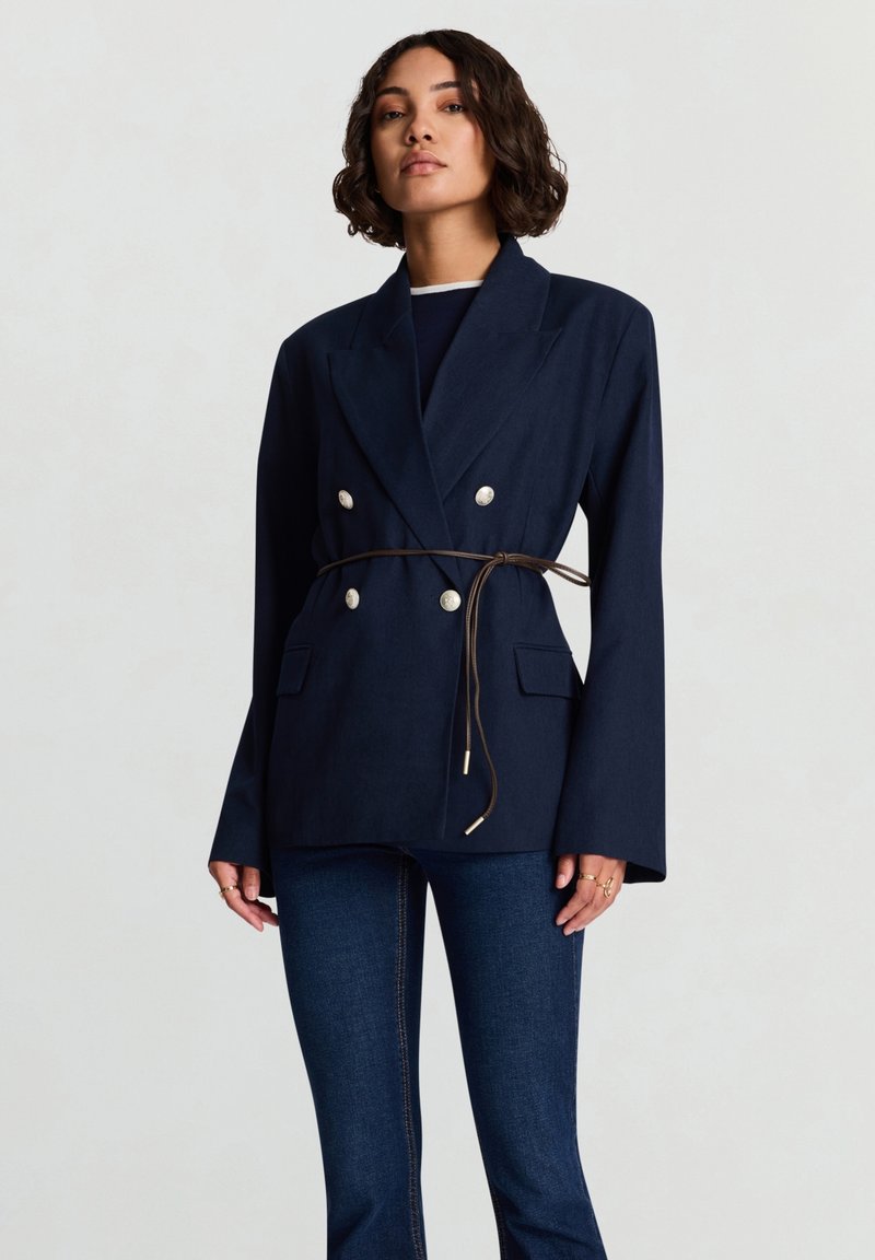 Blazer doppiopetto blu navy con cintura in vita, caratterizzato da due tasche frontali e grandi bottoni argentati; tessuto liscio con design strutturato.