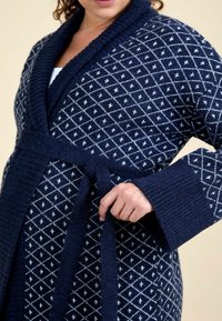 Femeie legând un brâu pe un cardigan tricotat bleu închis cu un model de diamante albe și cu manșete late cu nervuri peste un top alb.