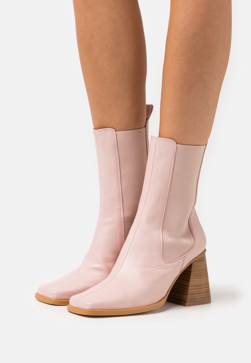 Personne portant des bottes en cuir rose pâle montant jusqu'au milieu du mollet, avec des bouts carrés et des talons carrés empilés en bois, debout contre un fond blanc.