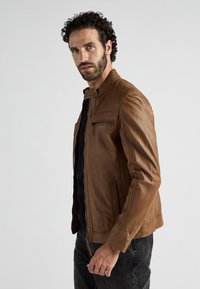 Mann mit lockigem Haar und Bart, der eine braune Lederjacke, ein schwarzes Shirt und dunkle Jeans trägt, steht vor einem schlichten hellen Hintergrund.