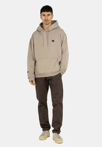 Beige Baumwoll-Hoodie mit frontaler Kängurutasche, verstellbarem Kordelzug und kleinem schwarzen Logo. Kombiniert mit braunen Hosen und beigen Sneakers.