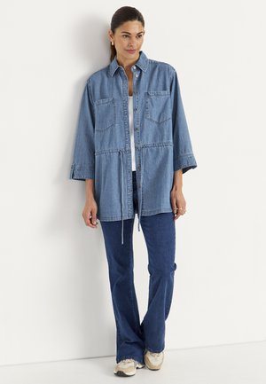 Kvinde iført en løs denimjakke over en hvid top, kombineret med mørkeblå, vidde jeans og lyse sneakers, stående op ad en hvid væg.