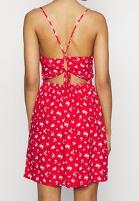 Mujer vestida con un vestido sin mangas rojo con estampado floral, con tirantes finos y un diseño de espalda abierta atada, de pie frente a un fondo liso.
