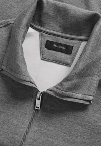 Grijze zip-up jas met een hoge kraag, gemaakt van zacht materiaal, met een zwart label met "Matinique" en een zilveren ritsdetail.