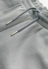 Pantalones de chándal gris claro con un cordón trenzado con puntas metálicas plateadas, cintura elástica y tejido suave y texturizado.