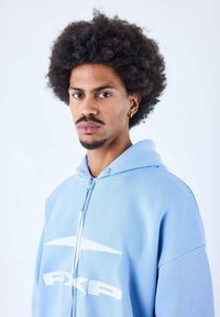 Sudadera azul clara de material suave, con cierre de cremallera, impresión de logo blanca y capucha con cordones. El modelo tiene el pelo rizado y lleva pendientes.