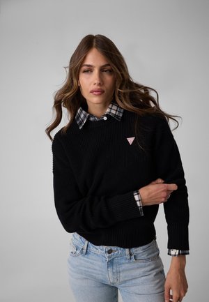 Kvinde med langt bølget hår bærer sort sweater over sort-hvid ternet skjorte og lyseblå jeans mod en ensfarvet grå baggrund.