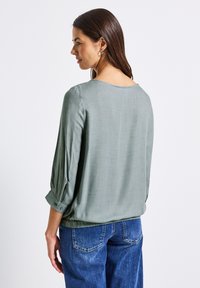 Blusa verde chiaro con maniche a tre quarti, texture morbida, scollatura sulla schiena a giro e polsini con bottoni, abbinata a jeans blu.