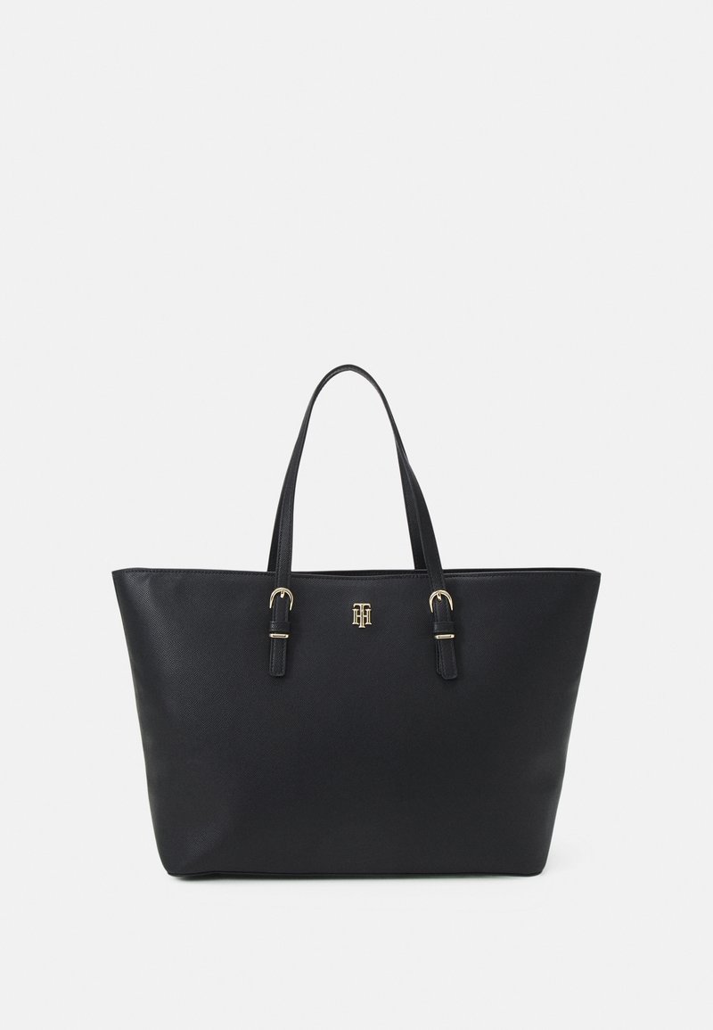 Sac cabas en cuir noir avec doubles anses et emblème logo doré centré sous les anses sur un fond clair uni.