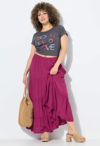 Graues, kurz geschnittenes T-Shirt mit mehrfarbigem Text, schichtiger fuchsiafarbener Maxirrock, gewebte hellbraune Tasche und hellbraune Absatzsandalen.