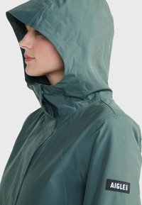 Veste verte imperméable avec capuche, présentant des lignes épurées, un col haut et un patch de marque sur la manche. Texture lisse, sans fermetures visibles.