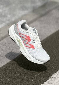 Bottine de course blanche avec une tige texturée, grand logo rouge, lacets fins et un accent jaune sur la semelle ; design léger et moderne.