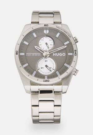 Reloj de acero inoxidable plateado con un dial texturizado en gris, tres subdiales, marcadores de hora en negrita y un taquímetro. Presenta una pulsera de eslabones de metal.