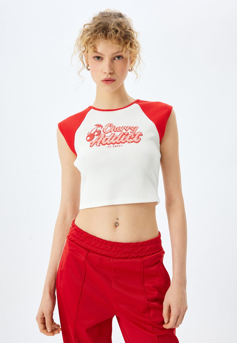 Top corto blanco con mangas en forma de gorra rojas, que presenta el texto "Cherry Addict" y gráficos de cerezas en rojo. Textura de punto suave. Pantalones rojos visibles.