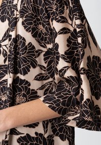 Svart blommönster på en beige tyg, med en avslappnad passform och vida ärmar. Slät textur med subtil lyster.