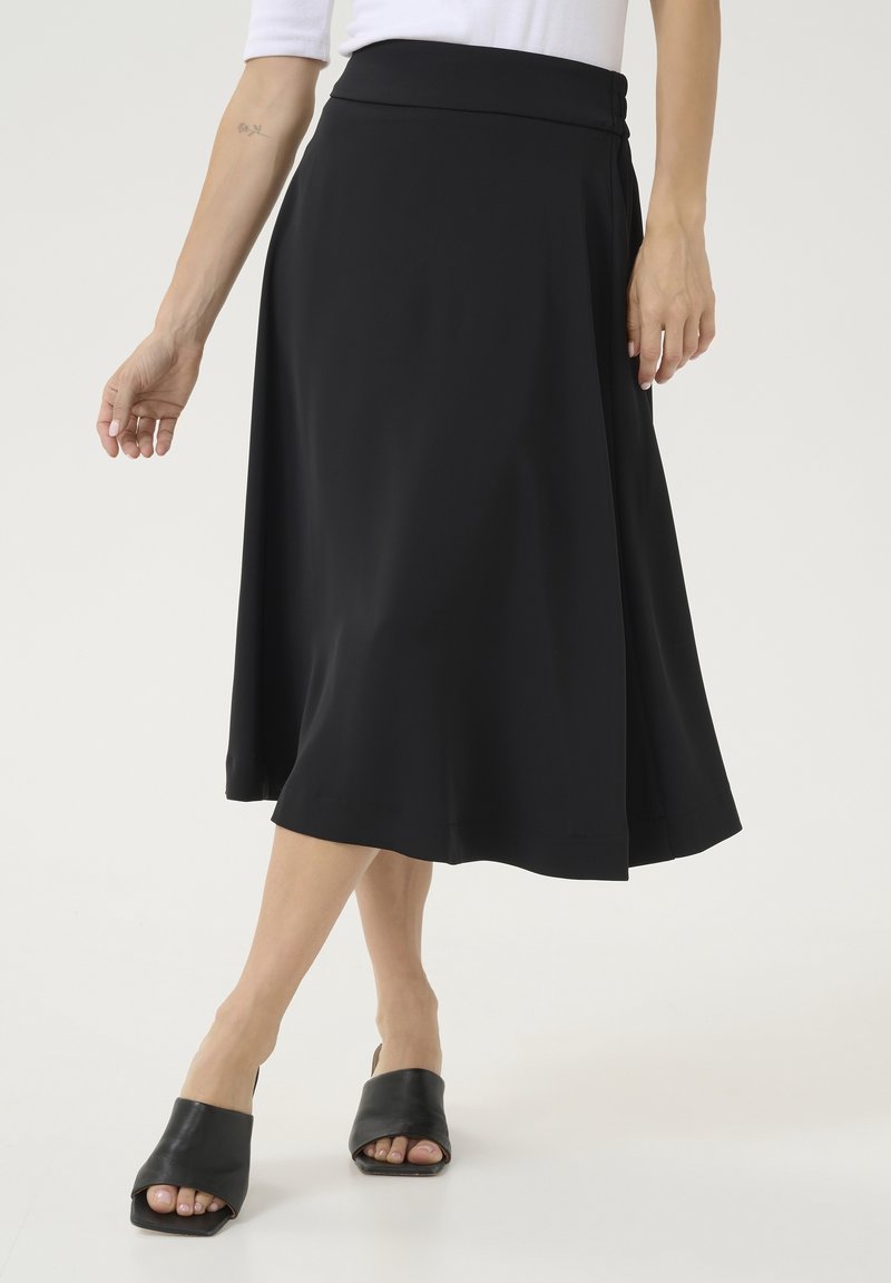 Femme portant une jupe midi noire, un haut blanc et des mules noires à bout ouvert, debout devant un fond blanc.