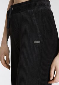 Schwarze Cord-Jogginghose mit vertikal geribbelter Textur, elastischem Bund mit Kordelzug und einem kleinen metallischen Logo-Highlight an der Seite.