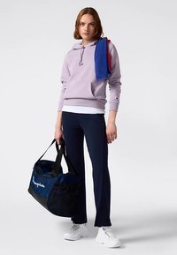 Lichtpaarse hoodie met voorzak, marineblauwe broek, witte sneakers, houdt een marineblauwe en zwarte duffeltas met het "Champion"-logo vast, blauwe en rode handdoek over de schouder.